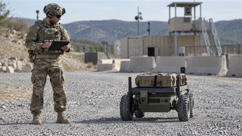 A soldier evaluates an unarmed logistics robot while using a rugged tablet at an outdoor test site.
